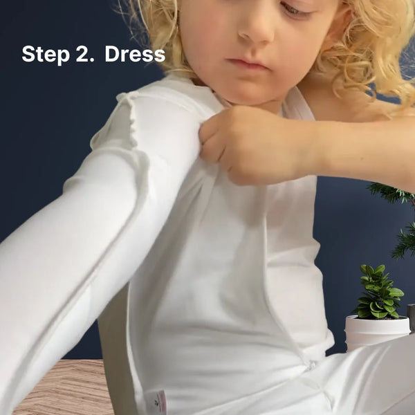Child getting dressed wearing a white skinakin suit with a plant in the background.